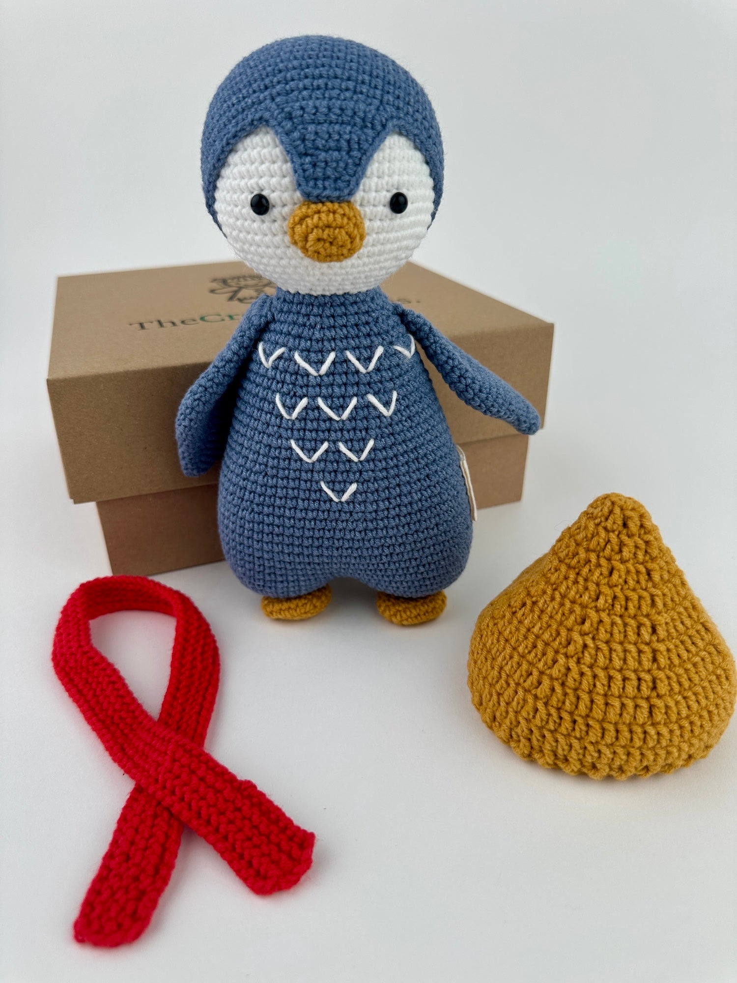 Crocheted penguin toy with a red ribbon and mustard yellow object on a white background